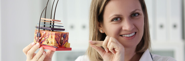 Smiling female dermatologist holding artificial human skin model and pointing at hair.