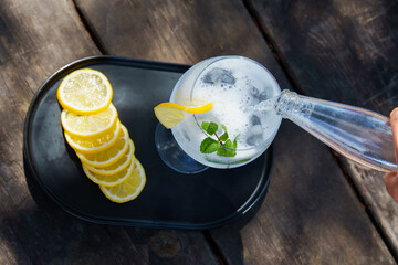 top view of a bottle of tonic poured over a glass with ice, slice of lemon and mint leaves,...