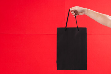 Hand of caucasian woman holding black gift bag with copy space on red background