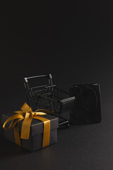 Vertical image of gift box with ribbon, gift bag, shopping trolley, copy space over black background