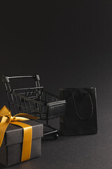 Vertical image of gift box with ribbon, gift bag, shopping trolley, copy space over black background
