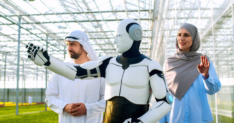 Robotic cyborg speaking with muslim man and woman about hydroponic system while walking at greenery. Concept of automated farming and artificial intelligence technology.