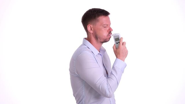 Young man checks blood alcohol level in light studio against white wall. Bearded male uses breathalyzer special device for alcohol level in blood