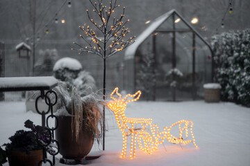 Snowy backyard with garlands and illuminated deer and glasshouse on background