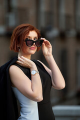 A confident and stylish redheaded woman poses for the camera, looking above her sunglasses with an...