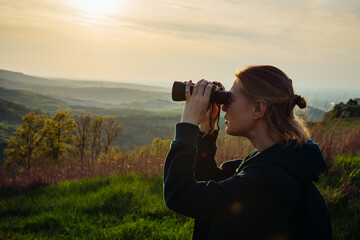 A hiker girl looks through binoculars at nature and birds standing on the top of a mountain in the bright rays of setting sun against the background of beautiful spring nature and cloudy blue sky - obrazy, fototapety, plakaty