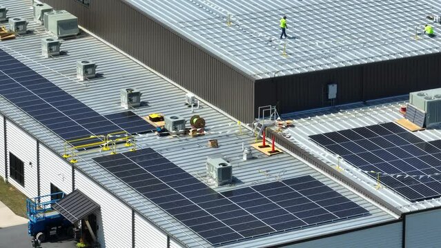 Industrial roof solar panel installation. Workers in neon on flat, metal rooftop preparing to install solar panel array. Aerial establishing shot.
