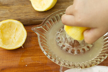 Little hand squeeze lemon juice with a lemon juicer glass on the wood table in the kitchen. Child...