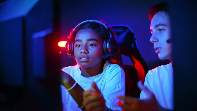 White boy and black girl teens in headsets discussing while playing video games in video game club with blue and red illumination. Keyboard and mouse with illumination