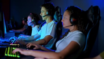 Multiracial group of teens in headsets playing video games in video game club with blue and red illumination. Keyboards and mice with illumination