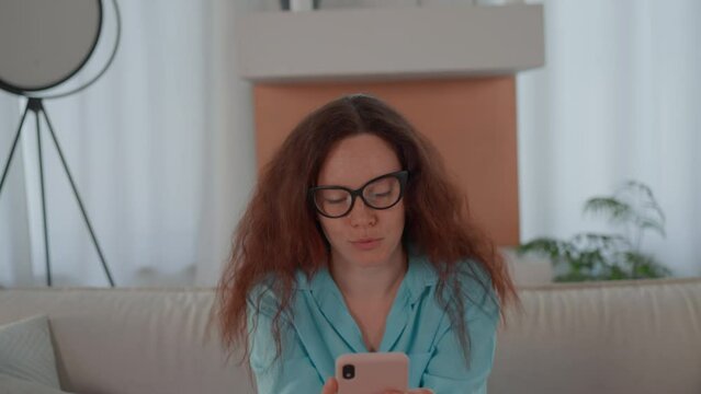 woman with red curly hair holding smartphone scrolling news feed showing different emotions sitting on the sofa in living room
