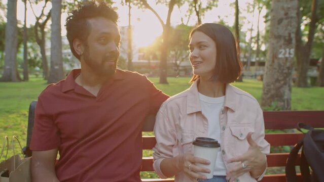 Medium arc shot of young diverse couple holding takeaway coffee cups and having talk while resting together on bench in park at sunset