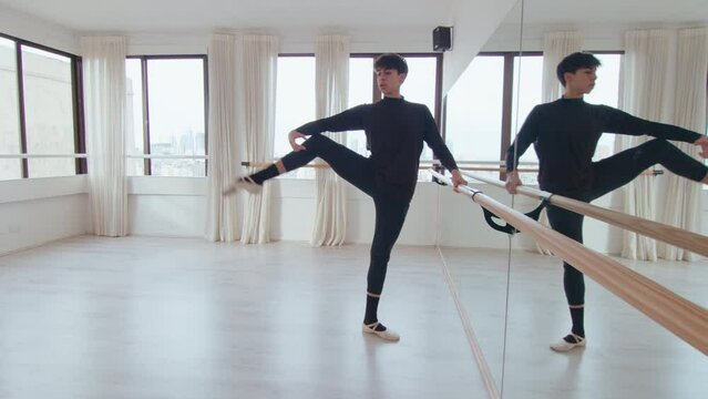 Young male ballet dancer performing passe, developpe and lunge at the barre in modern dance studio with mirror wall. Full shot with copy space