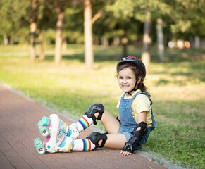 Cute little girl in protective equipment and rollers sitting on walkway in park outdoor. Summer activity for children.