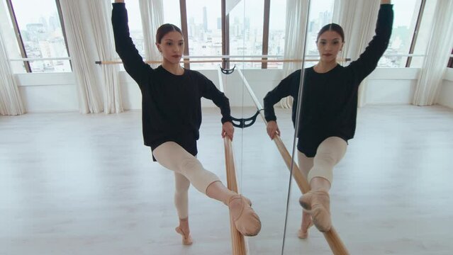 Young graceful ballerina holding one leg on barre, performing backbend and side stretch while exercising in a dance studio with a mirror wall. High angle view