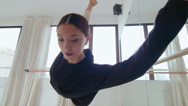 Young professional ballerina doing penche at the barre, leaning forward with one leg raised behind while exercising in dance studio. Zoom shot