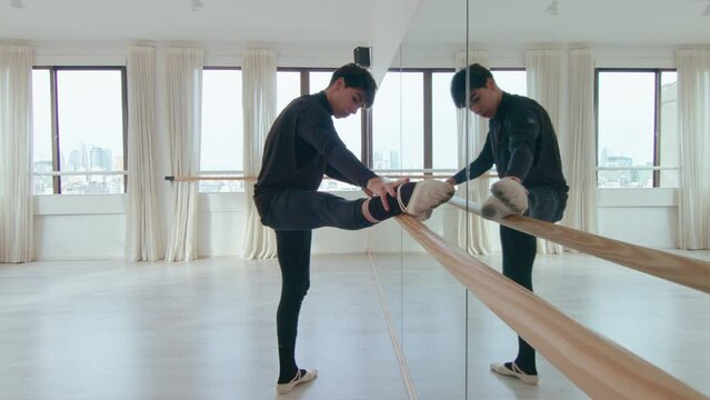 Young male ballet dancer holding one leg on the barre and stretching hamstring muscles while warming up in the studio. Full shot, zoom-out