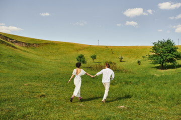 young couple of newlyweds running in green field, bride and groom in wedding gown holding hands
