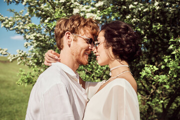 happy interracial couple in countryside, asian woman in white dress and man hugging near big tree