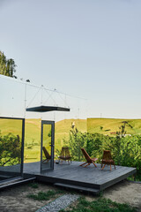 eco-friendly glass house with wooden porch near green hills under blue sky in scenic countryside