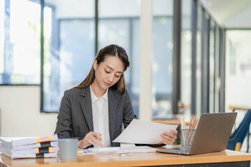 Asian businesswoman working with papers and laptop sitting at table in office, analyzing business accounting calculations and financial planning concepts. - obrazy, fototapety, plakaty