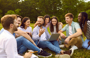 Group of happy, cheerful, diverse, multiethnic student friends sitting on green grass outside, studying information for project together, discussing something, using online tools on modern laptop PC
