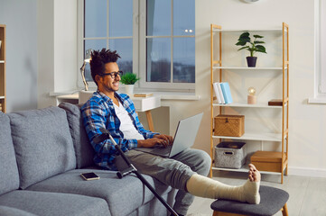 Young man with broken leg in plaster cast sitting on couch with laptop. Side view shot of smiling...