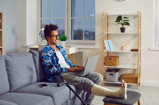 Young man with broken leg in plaster cast sitting on couch with laptop. Side view shot of smiling man in gypsum and crutch having online meeting. Rehabilitation, recovery and healthcare concept