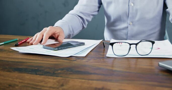 A man in a shirt works with financial papers, sitting at his desk, takes off his glasses and puts them on the table in front of him, the video camera moves in a circle.