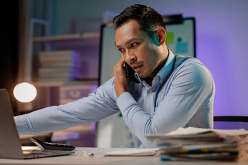 Asian man talking on the phone in the office of a startup company, company employees working overtime, late night work, overtime of a busy and hard working company employee. Overtime concept.