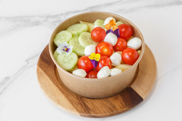 Salade composée de concombres, tomates, mozzarella et fleurs violette dans un bol en carton. Salade à emporter.