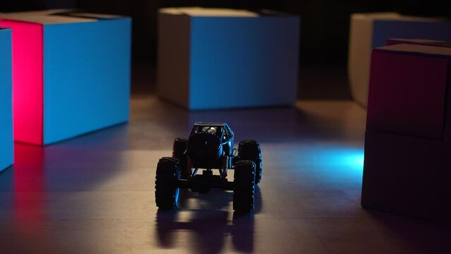 Radio-controlled toy car drives through improvised obstacles made of cardboard boxes on floor in dark living room on background of neon lights, no people, close-up, front view. Shooting in slow motion