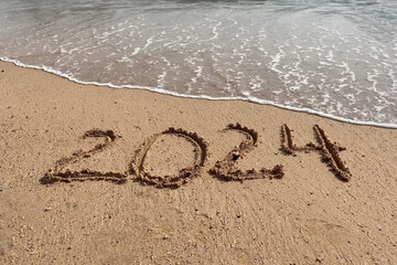 Happy New Year concept. Text 2024 handwritten in sand surface. Blue ocean wave washing away numbers on beach.