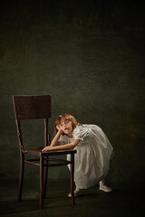 Portrait of boring little girl, child in white dress leaned on chair against vintage background. Fairy princess or queen. The heroine of story tale.