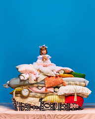 Portrait of smiling, happy little princess sitting on huge, big pile of pillows drink fresh, healthy cocktails over blue background. Fairy character.