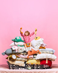 Portrait of pretty little princess sitting on big feather bed with pillows with retro phones over pink studio background.