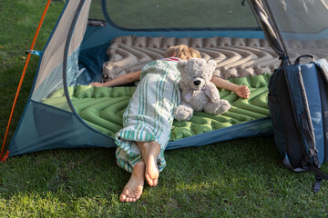 tired barefoot child sleeps or rests lying on his stomach in a tent with his favorite toy teddy...
