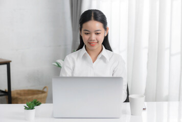 Asian young woman sitting at desk in office and drinking coffee, happy with the work they do on their tablet, laptop and taking notes at the modern office.