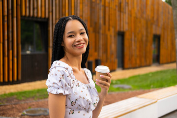 African young attractive woman in glasses stands on a bridge with a paper cup of coffee on the street during the day in the park in summer and looks at the camera