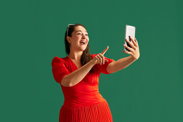 Pretty, smiling, happy woman in her 30s using mobile phone, taking selfie against green studio background. Social media. Concept of beauty, human emotions, lifestyle, fashion, ad