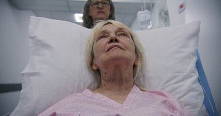 Fototapeta premium Senior woman lies on gurney, talks with mature female doctor. Physician transports patient to hospital ward down modern medical center hallway after surgery. Medical personnel at work in hospital.