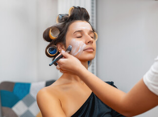 Fototapeta premium make up artist applying powder,cream, foundation, painting eyebrows, lips,putting false eyelashes.woman with curlers on hair. ready girl posing against wall,fingers on lips.brushes in pants pocket