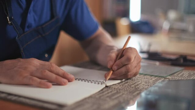 Medium close up of left-handed man drawing sketch with pencil in his notepad while sitting at table in workshop