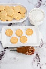 ingredients and mise en place for the preparation of cornstarch alfajores, filling the dulce de leche filling on top and grated coconut on the outside on white plates.