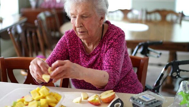 Elderly senior woman cuts up fruit with arthritis hands and she struggles to cut up an apple fruit with a knife.