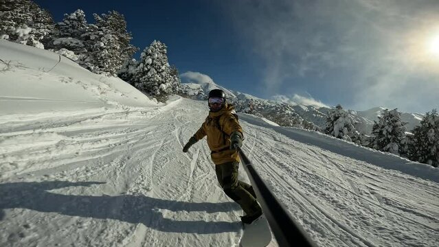 Man riding on snowboard with selfie stick in his hand between trees on slow motion. Guy doing snow splash rising on magnificent sunny day. Concept of extreme in sport - winter, freeride, snowboarding