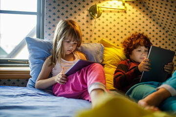 Young brother and sister playing video games together on their tablets in a bedroom