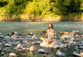 Beautiful young woman sitting alone on stone in the mountain river with closed eyes and hand on chest breathing and  enjoying the silence and beauty of nature at sunset. Mindfulness, spirituality 