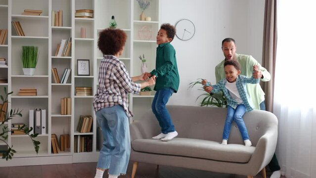 Multiracial family of mother, father and two preschool boys fooling around and spending active free time at home. Energetic kids holding parents' hands and jumping on couch. Leisure activity indoors.