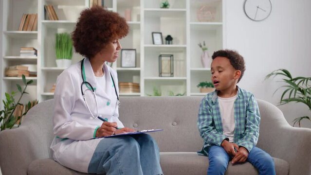 Caring female baby doctor having appointment with cute little boy and conducting examination in medical office. Smiling woman checking kid's pronunciation and showing okay gesture.